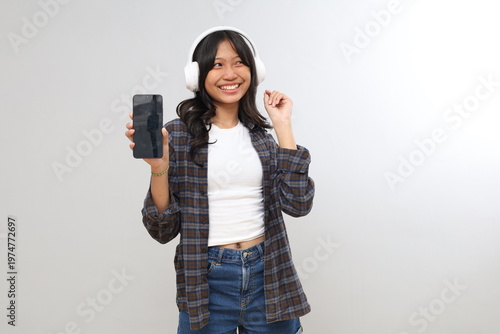 Schilderij op canvas A young Asian teenage girl shows a blank screen of her smartphone while listening to music through headphones, isolated on a gray background