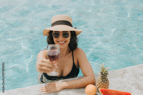 Young beautiful woman in black bikini wearing hat and sunglasses relaxing in swimming pool. Caucasian female in swimwear chilling in pool. Summer vacation concept.