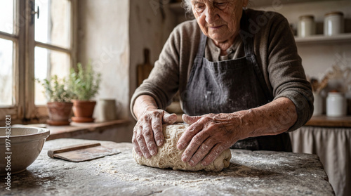 Elderly woman prepares dough in a rustic kitchen while using her hands and a stone surface