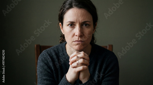 Woman sitting at a table with hands clasped, looking directly at the camera while showing an expression of concern in a simple room setting