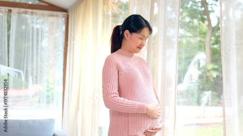Happy asian pregnant woman wearing pink sweater standing near bright window gently holding large belly expressing peaceful maternity moments expecting beautiful healthy newborn baby in cozy home