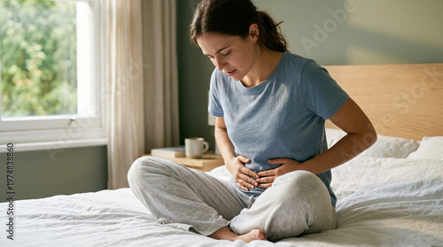 Young woman sitting on bed feeling intense stomach pain holding abdomen suffering from severe cramps indigestion or menstrual ache expressing discomfort and distress in bright cozy bedroom