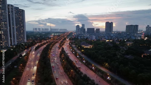 Drone view of curved boulevard in Hanoi with steady traffic, surrounding greenery, and residential towers under soft evening sky near sunset