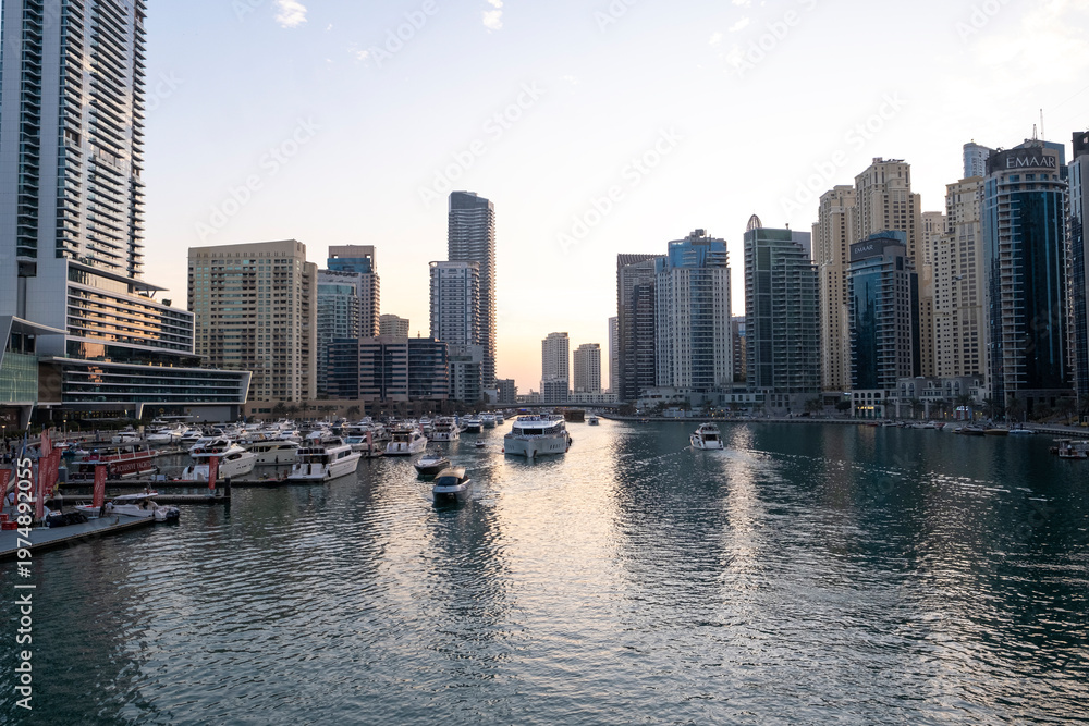 Obraz premium 12.12.2025 Dubai, UAE. Luxury yachts and boats moored in Dubai Marina at sunset. Stunning view of skyscrapers and modern architecture in UAE with golden sky reflection in the water. High-end lifestyle