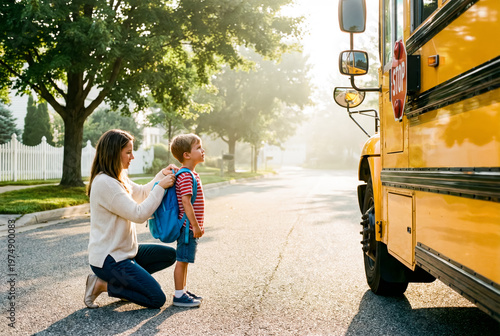 Mother helps her young son put on a backpack next to a yellow school bus.