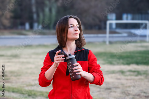 Fit mid adult brunette woman in her 40s drinking water from a sports bottle after outdoor stadium workout