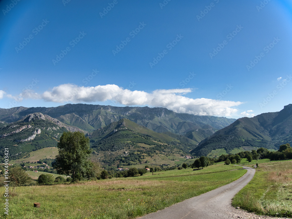 Fototapeta premium Winding Road Through Mountain Valley in Asturias - Scenic view of a paved road cutting through lush green meadows with rolling mountains under a bright blue sky.