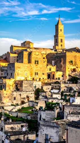A sun-drenched, vertical view captures a historic town. Buildings cascade down a hillside, crowned by a prominent bell tower under a blue sky