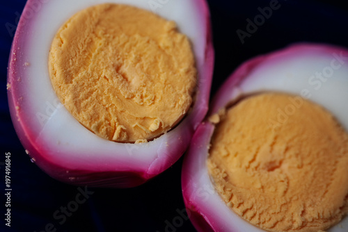 Macro close-up of two halved pickled eggs with vivid pink outer whites and firm golden yolks on a dark plate. High detail texture with minimal background.