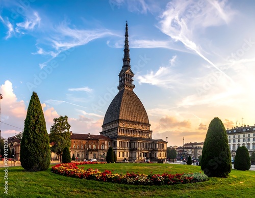 A tall, stone building with a spire dominates a public park. The sky is bright, and the sun sets in the background