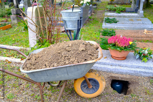 Dark funeral urn placed in stone grave niche beside rustic wheelbarrow with fresh earth.  Geneva. Switzerland.