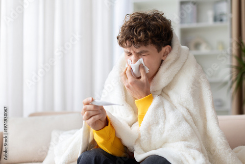 Fotografía Sick teen boy wrapped in white blanket blowing nose with tissue holding thermometer on sofa