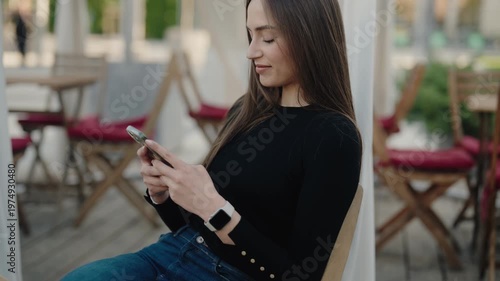 Young woman in casual attire sitting outdoors, happily using her smartphone. Relaxed and positive atmosphere in a casual setting. Young Woman Texting on Smartphone in Outdoor Cafe Setting