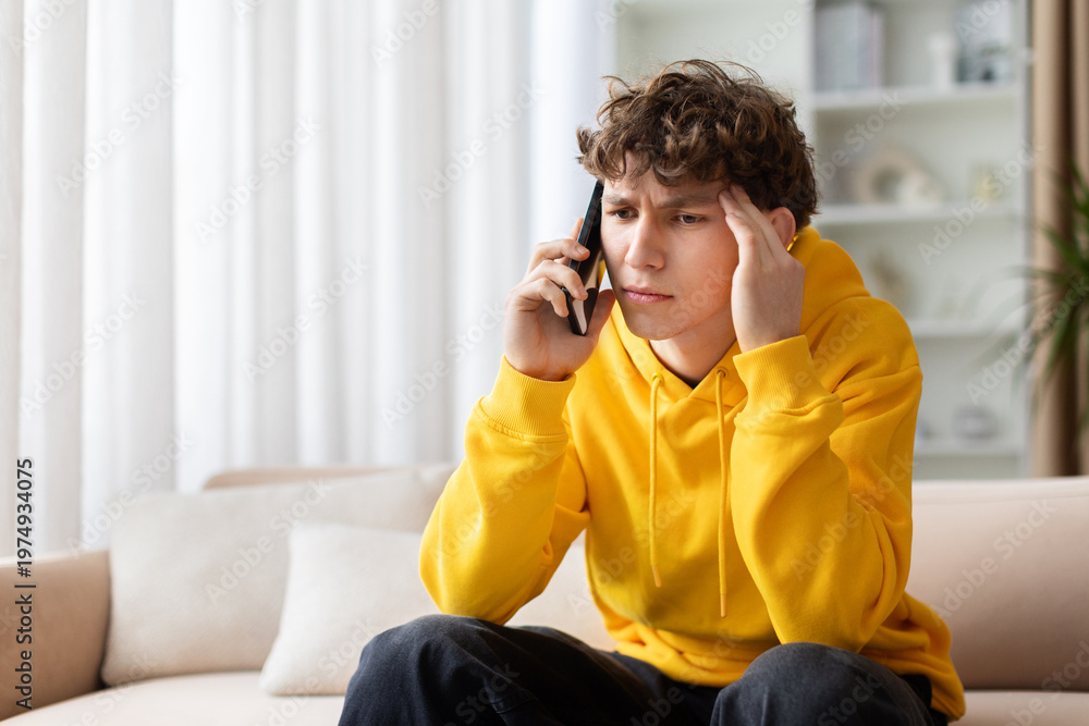 Obraz premium Worried teen boy in yellow hoodie talking on phone with hand on temple, looking stressed. Young troubled male having difficult call at home on sofa