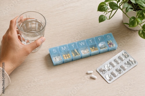 hand holds glass of water near weekly pill organizer and blister pack