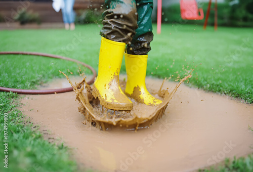 Closeup child feet in yellow rain boots create a massive splash as jump in a muddy puddle. Water ripples outward as the kid play into the mess on a green lawn of the backyard. Carefree childhood joy