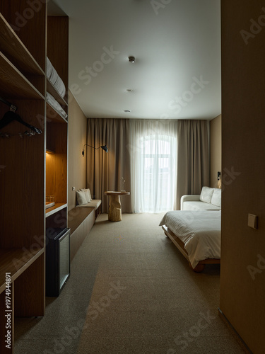 Modern hotel room interior with wooden wardrobe, long cabinet, and twin beds near a large window with sheer curtains. Warm beige tones and minimalist design.