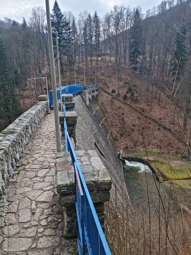 Stone dam in Międzygórze in a mountain valley, peaceful landscape with river and bridge, combination of nature and hydraulic engineering.