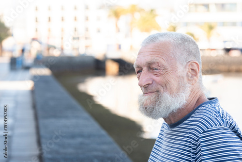 Εκτύπωση καμβά Elderly bearded man smiling in profile on a sunny seaside promenade wearing a st