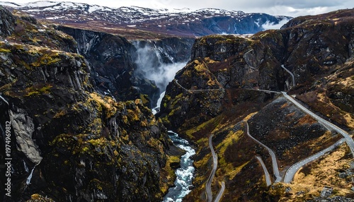 Scenic gorge view with rushing river, cascading waterfall, winding roads, and distant snow-capped mountains on overcast day