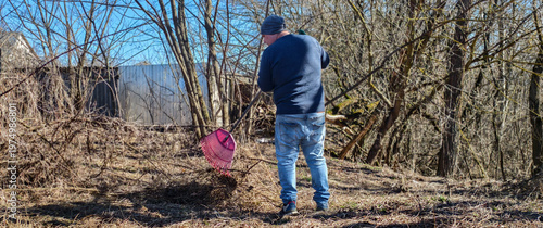 Photography Man wearing jeans and a blue sweater raking dry leaves and branches in a neglected backyard during a sunny early spring day, engaging in garden cleanup and yard work