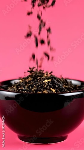Dried tea leaves are falling into a black ceramic bowl against a vibrant pink background