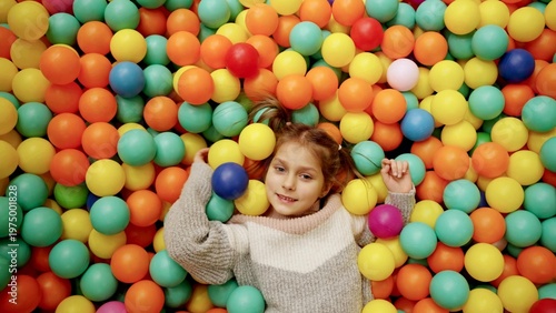 Фотография Happy little joyful cute girl playing with colorful balls in playground ball pool, lying on the floor in game center