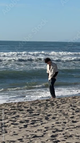 Teenager walking along a sandy beach, gazing towards the ocean, with waves crashing in the background. Reflective and tranquil moment captured during sunrise at the coastline.