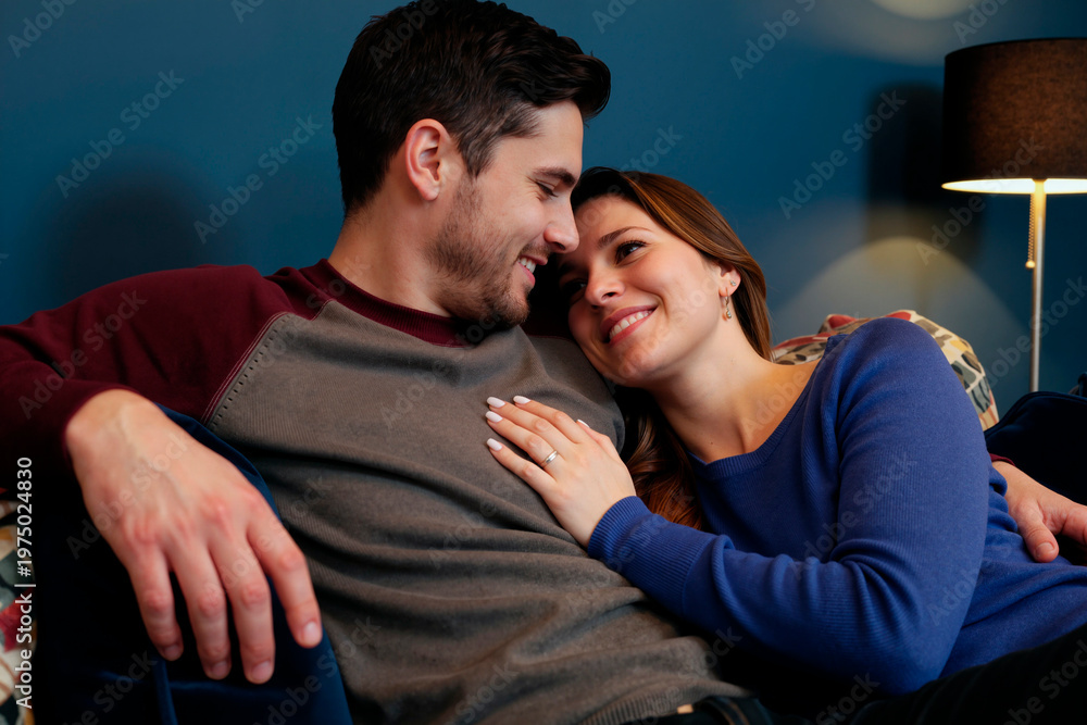 Obraz premium Caucasian young adult couple cuddling on sofa, smiling and leaning together, expressing intimacy and relationship trust. Ideal for dating, marriage, home life, or Valentine marketing