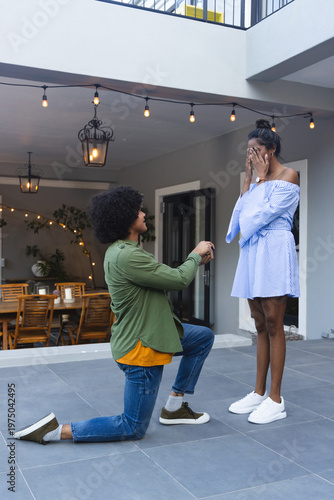 African American couple kneeling and proposing on tiled patio under string lights, ring box visible