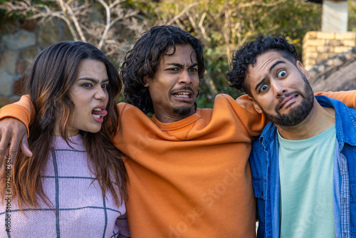 Diverse friends wearing pink sweater, denim shirt, making exaggerated faces in yard near stone wall
