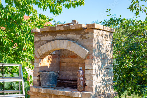A large outdoor brick fireplace stands completed in a sunny green garden. outdoor cooking, backyard construction, stone oven.