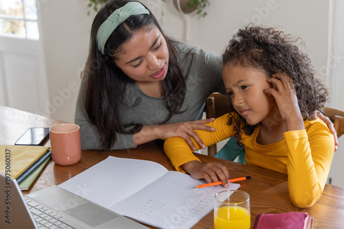 Woman and child leaning over notebook at dining table, pointing at math with orange pen