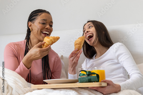 Diverse women sharing breakfast in bed holding croissants and tray with orange juice and gift box