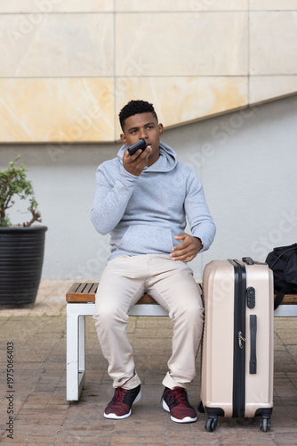 African American man sitting on bench talking on smartphone, holding beige suitcase, dark bag