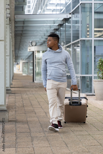 African American man walking under metal canopy along tiled walkway pulling tan rolling suitcase