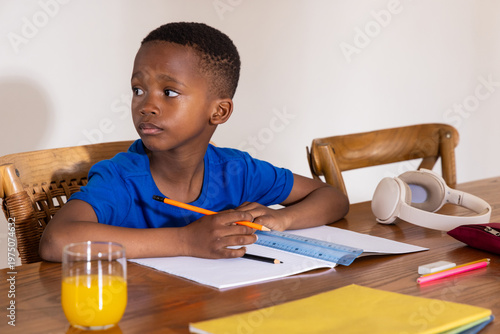 African American male child sitting at wooden table in blue tee, holding pencil, headphones nearby