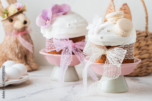 Easter cakes decorated with white cream, macaroons and edible violet flowers. Traditional sweet Easter bread on the stand with colorful painted eggs and straw bunny. Easter themed background