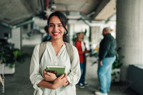 Young female university student smiling, holding books and backpack, standing in a college campus during a break time