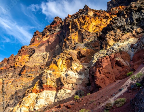 A vibrant shot of a mountain range displaying unique geological formations. The layers showcase shades of orange, red, black, and white under a partly cloudy blue sky