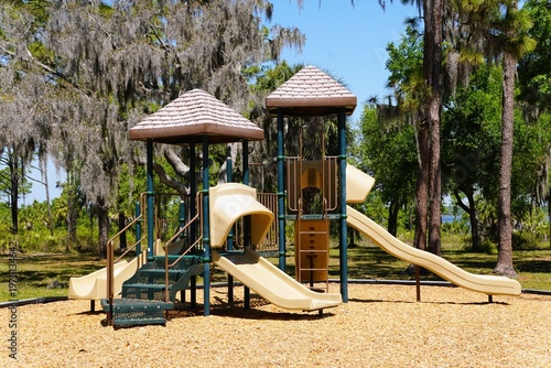 A playground with two roofed towers, slides, stairs, and climbing features stands among tall trees with hanging moss near Lake Manatee State Park, Bradenton, Florida, U.S