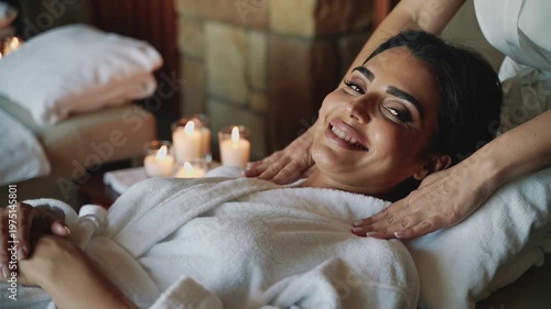 A serene woman lies comfortably on a massage table, eyes closed, as skilled hands gently massage her head and body, surrounded by the soft glow of can