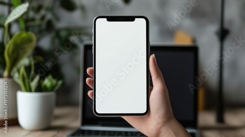 Female hand holding smartphone with blank screen in front of laptop