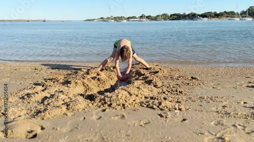 Young child enthusiastically digging a long trench in the wet sand at the water's edge on a beautiful sunny day, with sailboats visible on the blue water in the background