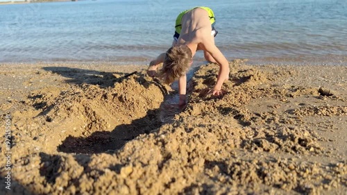Energetic boy in swim trunks happily digging a channel in the wet sand to let the ocean water flow, enjoying his summer vacation playing on the sunny shoreline during a beautiful sunset