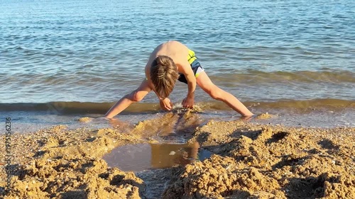 Happy child playing with wet sand on the seashore, building a small dam and a channel to direct the gentle waves of the sea during a sunny summer day on vacation at the seaside resort