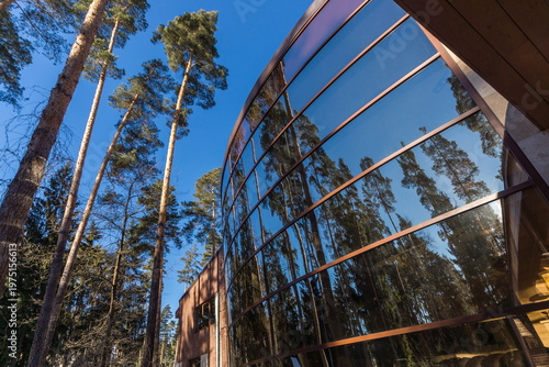 A large building made of glass and wood stands beside tall trees in a forest. The glass reflects the green surroundings. The sky is clear and blue