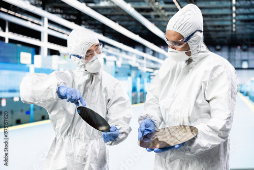 Photos Man and coworker in microchip industrial plant comparing silicon wafers, wearing bunny suit