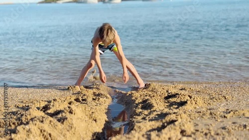 Cheerful young boy playing in the sand at the seashore, digging a trench with his hands to let the seawater flow, enjoying a sunny day during his summer vacation by the beautiful blue ocean