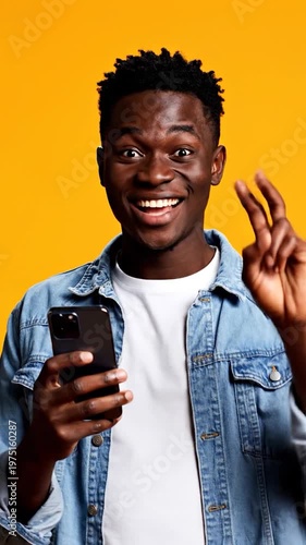 A happy young man in a denim jacket looks at his smartphone while standing against a bright yellow background